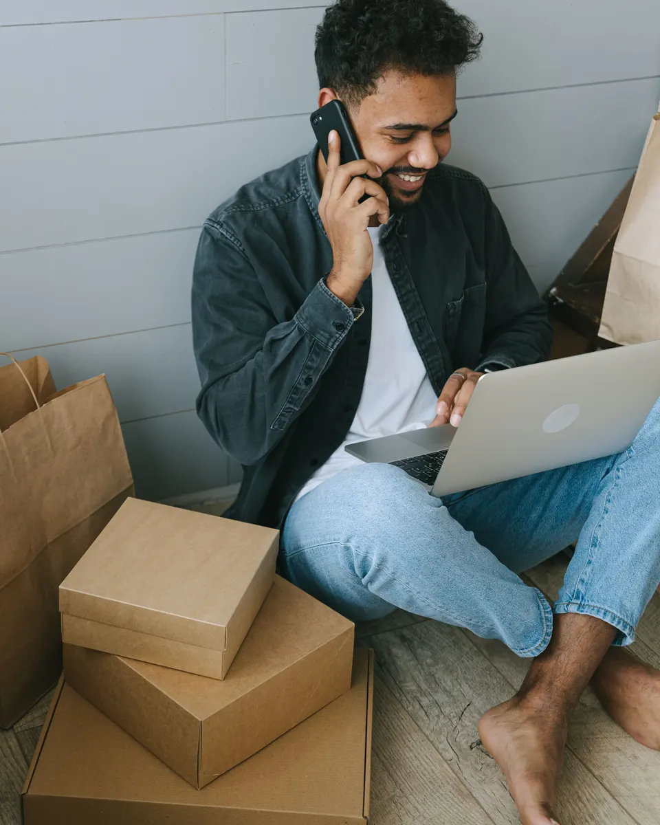 A smiling man on the phone with a laptop, beside stacked parcels